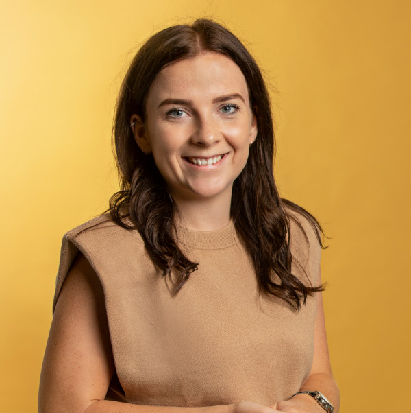 Een vrouw in een lichtbruine jurk staat glimlachend voor een gele achtergrond ingehuurd door Hypotheken in Almere.