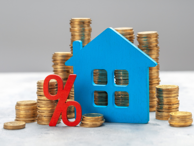 Een blauw houten huis en een rood percentagesymbool worden omringd door stapels munten. Deze munten vertegenwoordigen een investering in onroerend goed of een hypotheekrente.