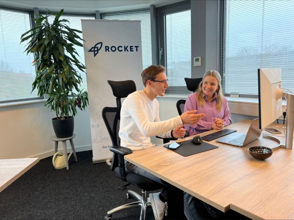 Twee mensen zitten aan een bureau in een kantooromgeving, verwikkeld in een discussie. Een "Rocket"-banner staat op de achtergrond, samen met een potplant en een raam met jaloezieën.