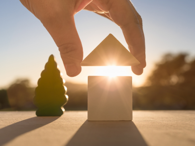 Een hand plaatst een houten driehoekig blok op een vierkant blok, dat lijkt op een huis, met de zon erdoorheen schijnend. Een boomvormig blok is links.